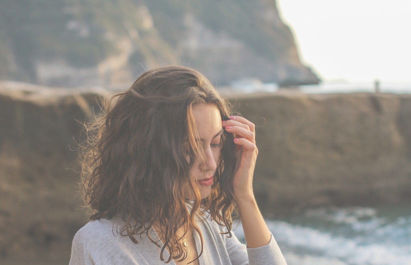 Photograph of a woman with wavy brown hair wearing a gray sweater, standing near a rocky shoreline with waves and a large hill in the background. The scene captures a contemplative mood with natural lighting and muted colors.